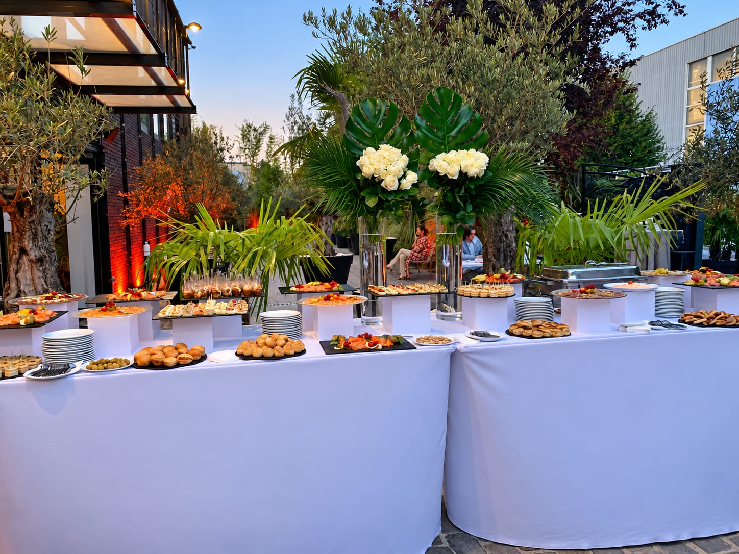 Buffet de cocktail dressé au jardin à la tombée du jour, sous des bouquets de roses blanches