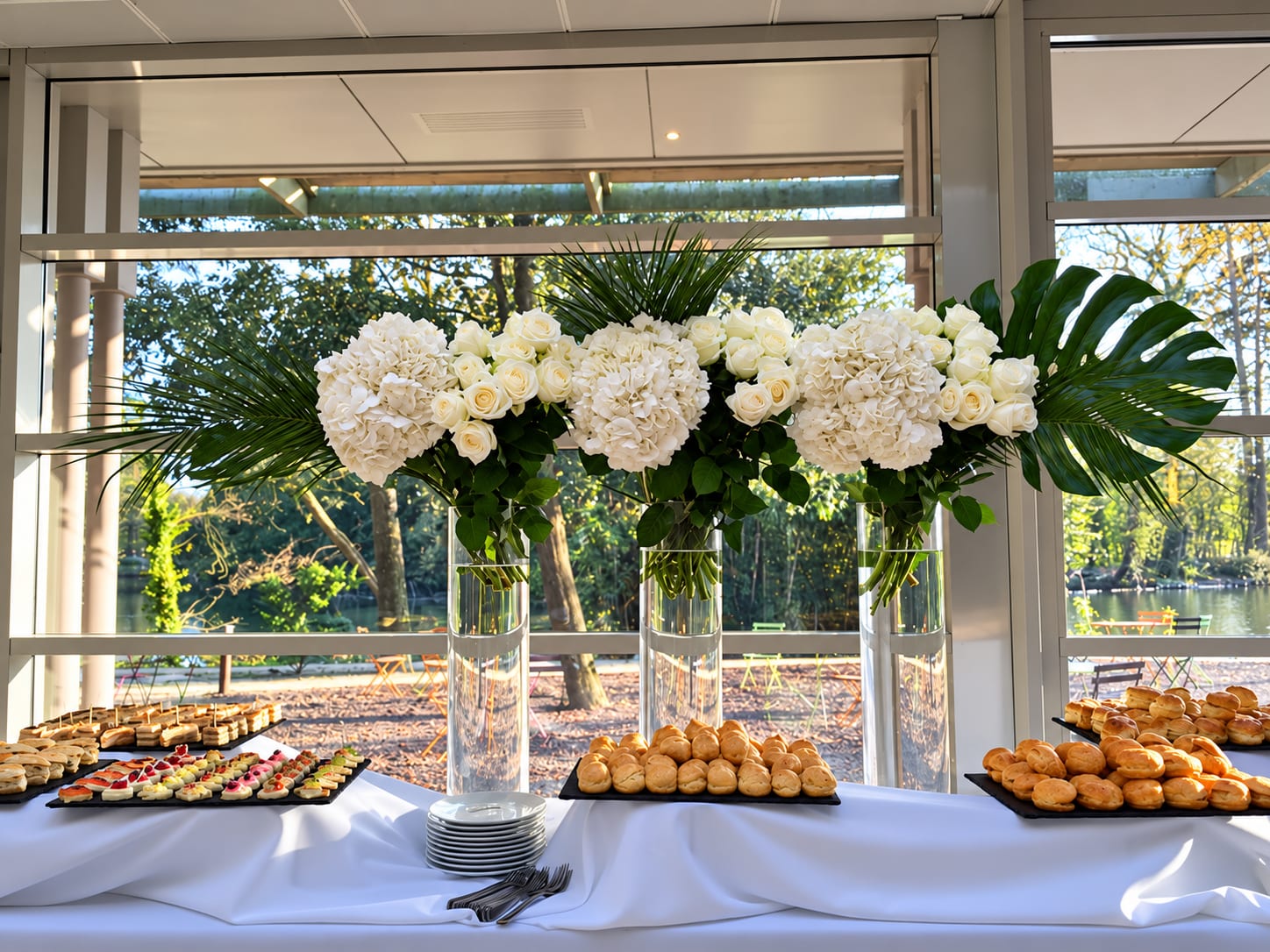 Hortensias et roses blancs en vases hauts, palmes et monstera, au-dessus des canapés et des gougères, devant les baies vitrées sur le lac