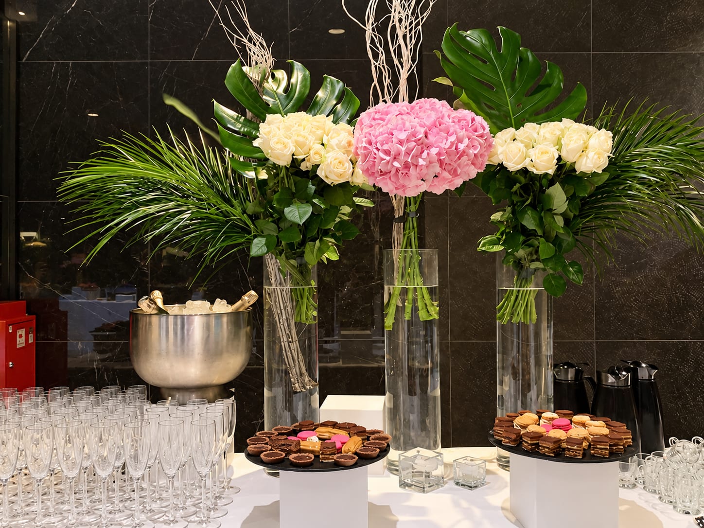 Trois bouquets de roses blanches et d’hortensia rose, monstera et palmes, devant un mur de marbre noir, flûtes de champagne et mignardises