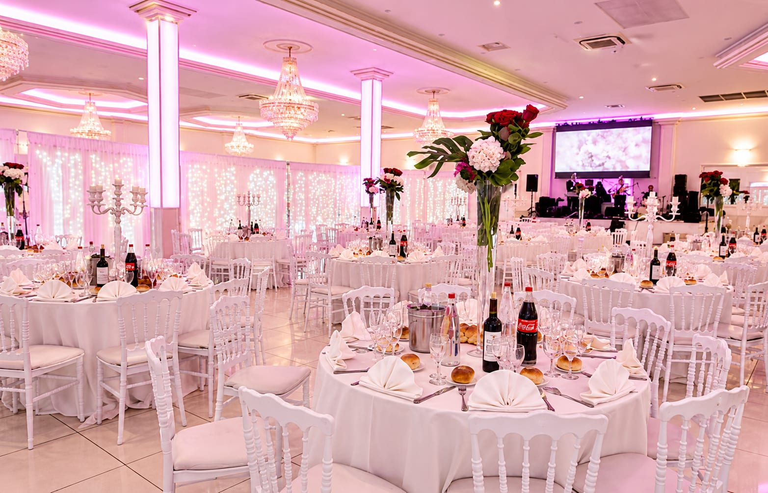 Salle de réception dressée de tables rondes, chacune sous un grand vase de roses et d’hortensias