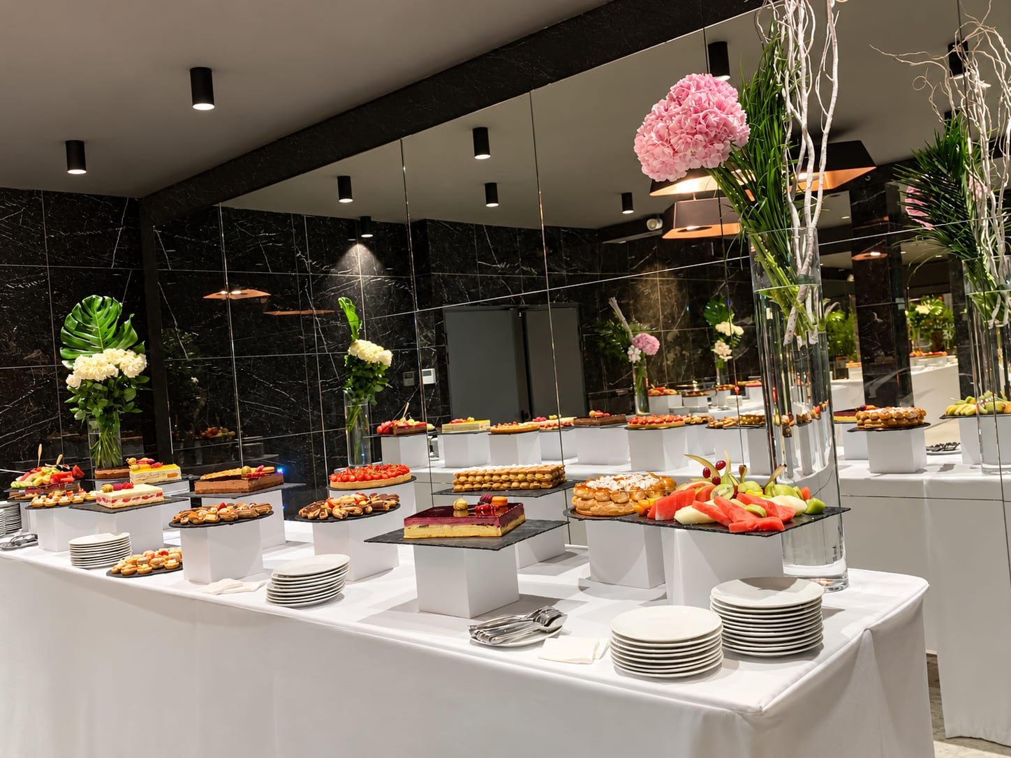 Buffet de desserts dans une salle aux murs de marbre noir : tartes, entremets et fruits sur présentoirs, fleurs blanches et roses